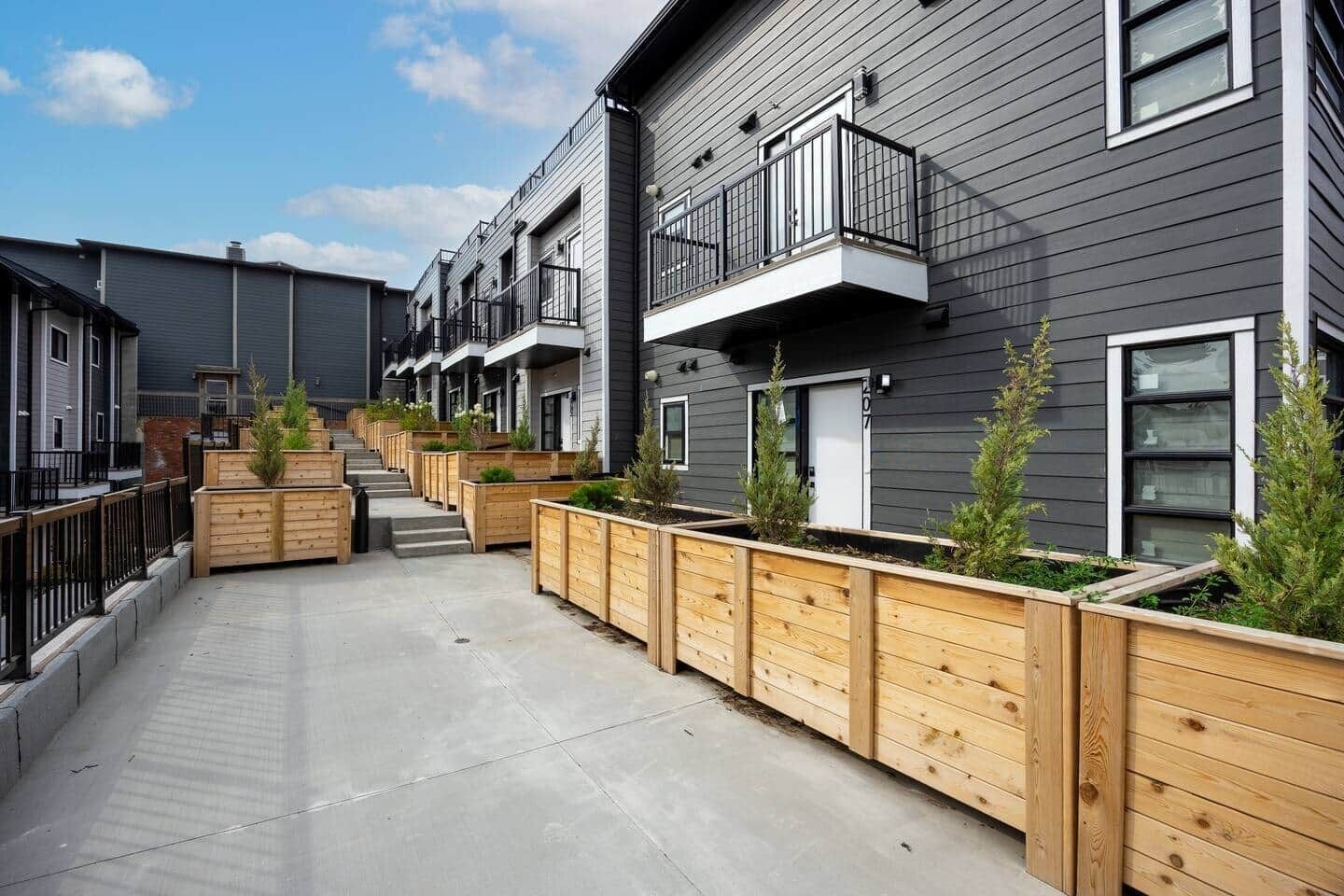 A modern residential building with multiple balconies, wooden planters with greenery, and a concrete pathway.