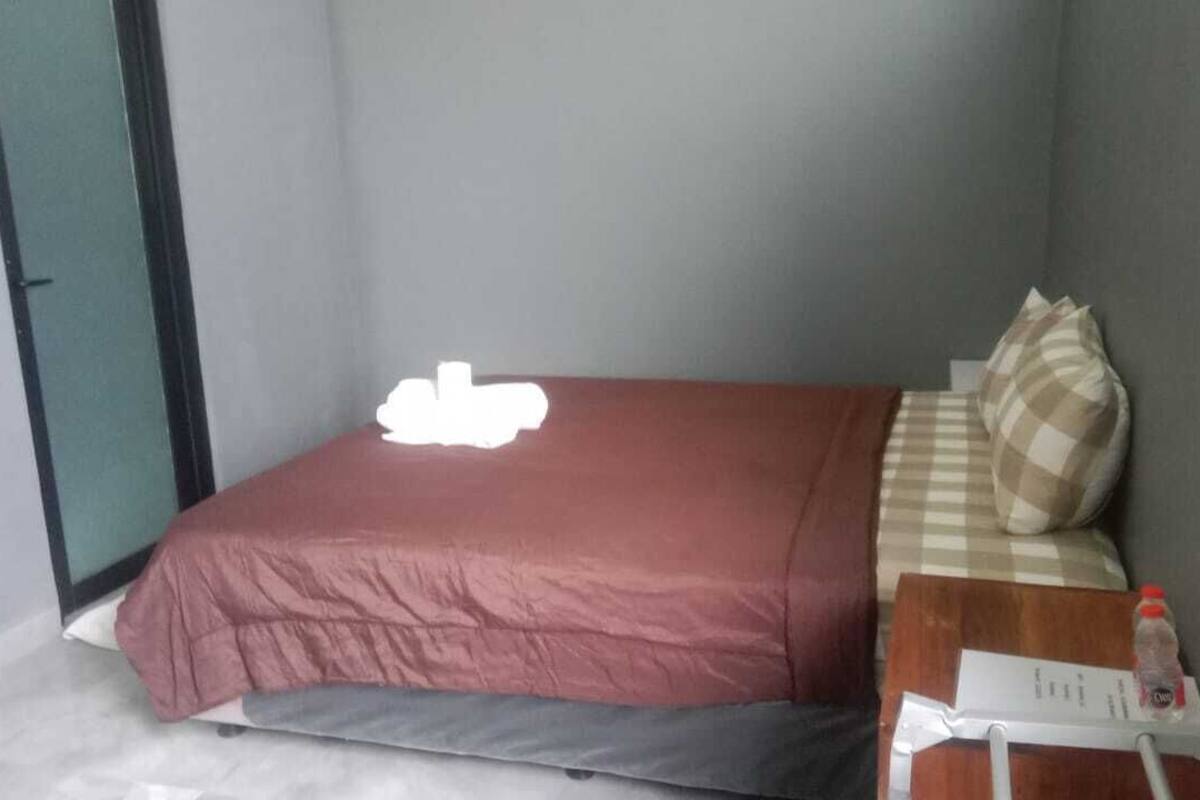 Una cama pequeña y ordenada con colcha roja y almohada a cuadros. Una botella de agua y un papel están sobre la mesita de noche.