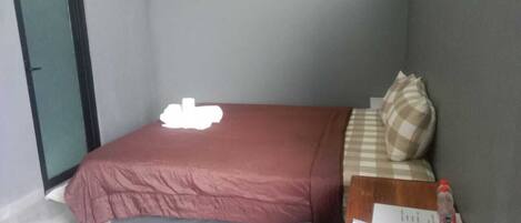 A small, neatly made bed with a red bedspread and a checkered pillow. A water bottle and a piece of paper are on the side of the bed.