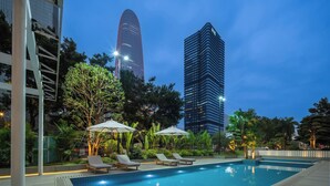 A swimming pool with lounge chairs and umbrellas, surrounded by greenery and tall buildings.