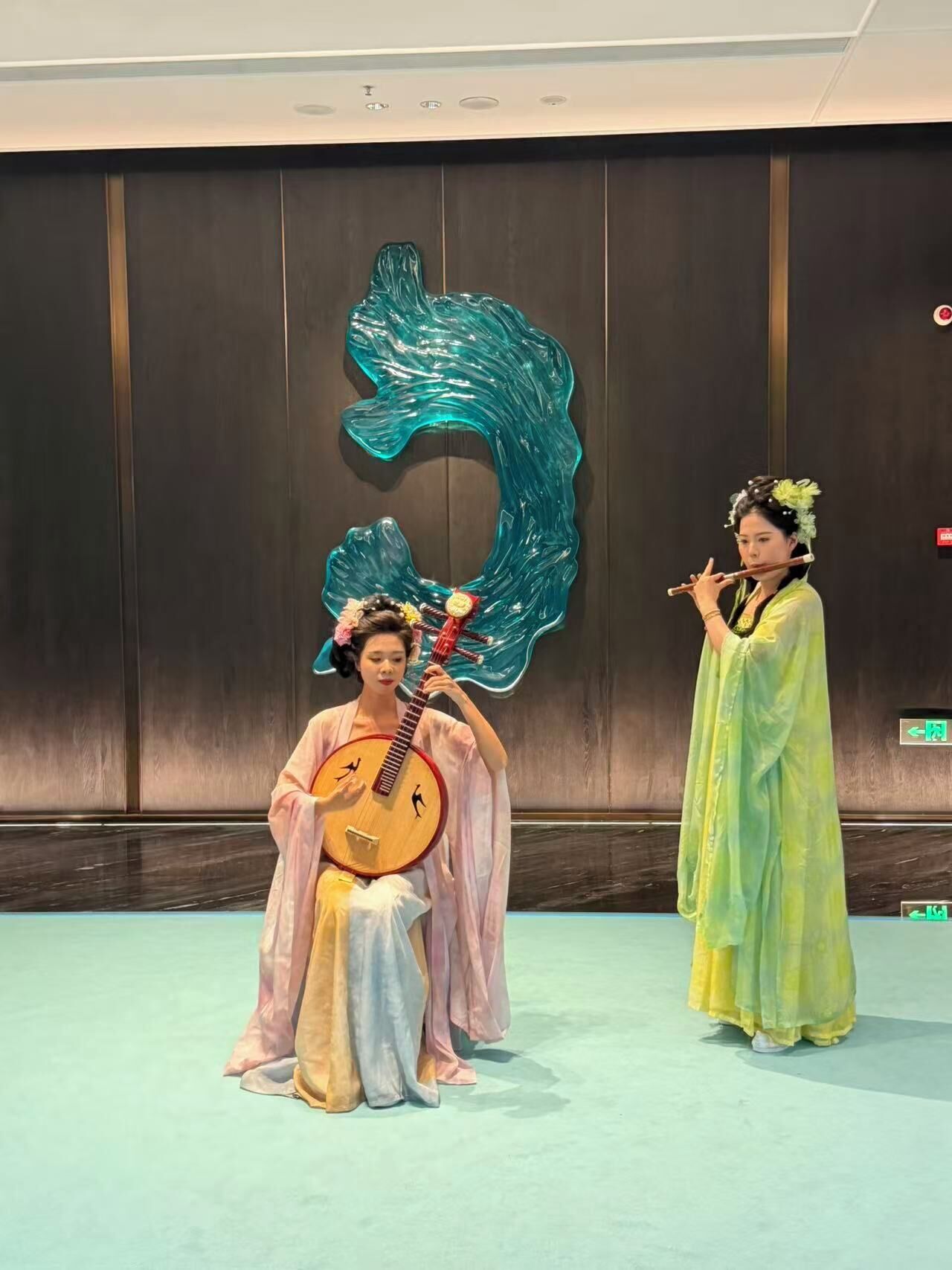 Two individuals in traditional attire, one playing a stringed instrument and the other holding a flute, in a room with a large decorative wall piece and a light blue floor.
