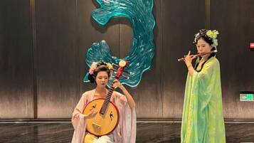 Two individuals in traditional attire, one playing a stringed instrument and the other holding a flute, in a room with a large decorative wall piece and a light blue floor.