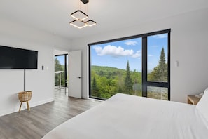 A modern bedroom with a large bed, a TV mounted on the wall, and a view of a forest through large windows. - The Apex: Prestigious Tremblant Experience (Mont-Tremblant)