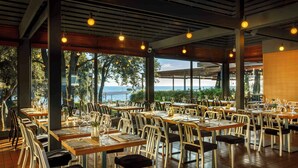 Ein geräumiges Restaurant mit mehreren Tischen und Stühlen, große Fenster mit Meerblick und warmem Licht.