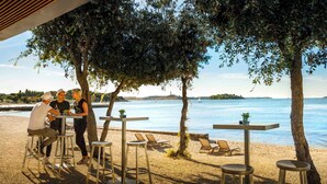 Ein Café direkt am Strand mit Terrassenplätzen, mit Blick auf ruhiges Meer und eine entfernte Küstenlinie.