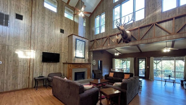 A spacious living room with wooden walls, a fireplace, and a mounted deer head. - Spacious Lodge Retreat | Sleeps 24 | 8 Suites | Decks, Pond Views & Gathering Spaces (Zwolle)