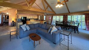 A spacious living room with a large sectional sofa, a coffee table, and a dining area with chairs and a table. A kitchen with wooden cabinets and a refrigerator is visible in the background. - Relax & Unwind in this Vaulted-Ceiling Cabin with Fireplace, Full Kitchen & Lake Views (Zwolle)