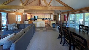 A spacious living area with a sofa, dining table, and chairs, a kitchen with wooden cabinets, and large windows offering views of the outdoors. - Relax & Unwind in this Vaulted-Ceiling Cabin with Fireplace, Full Kitchen & Lake Views (Zwolle)
