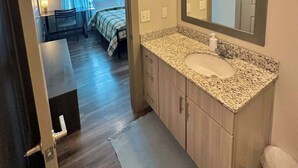 A bathroom with a granite countertop, a sink, a mirror, and a door leading to a bedroom with a bed and curtains. - Luxury Midtown Apt: Pool, Gym & 4 Bedrooms! (Atlanta)
