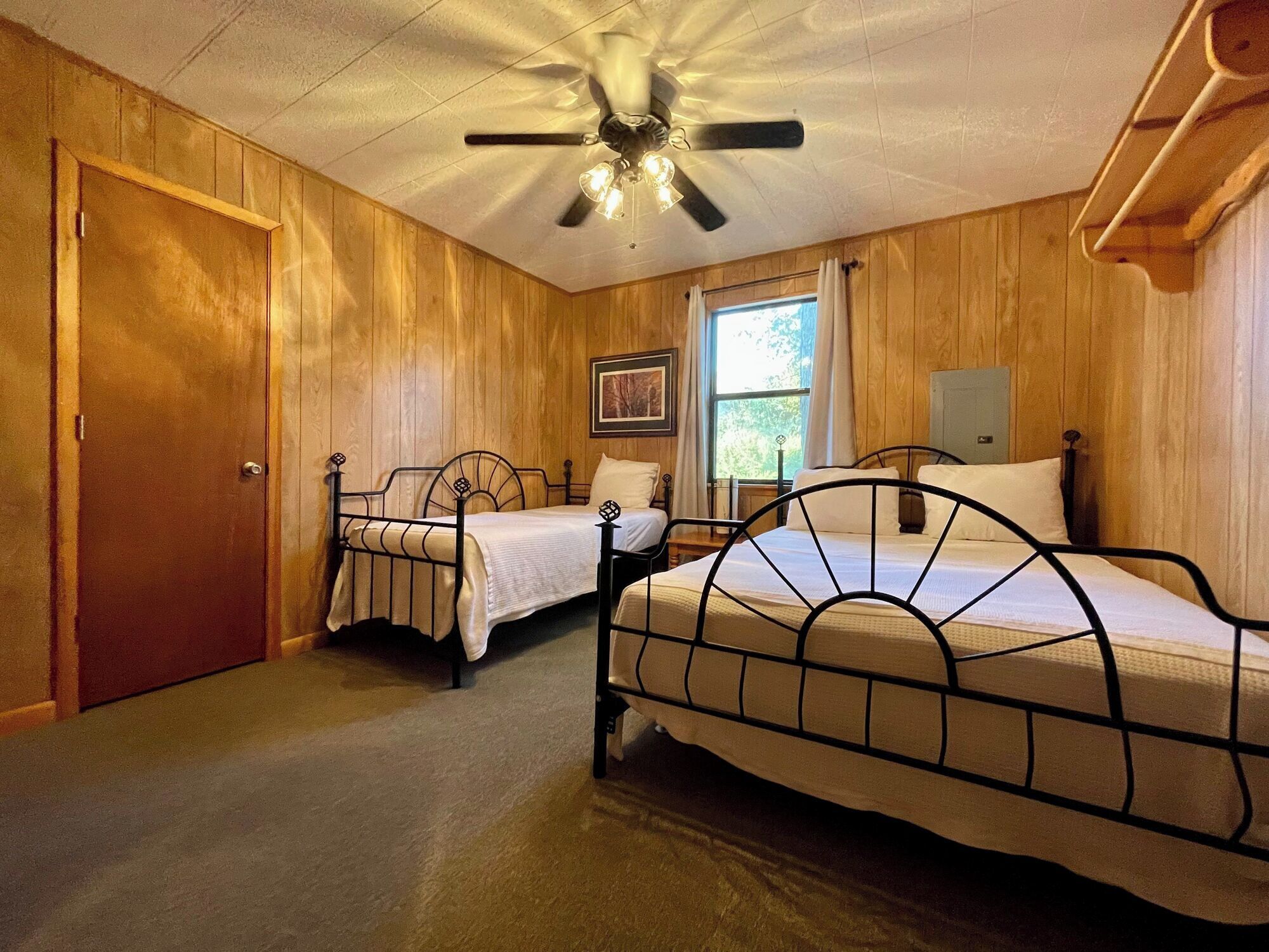 Una habitación con paneles de madera, dos camas de metal, un ventilador de techo, una ventana con cortinas y una puerta.