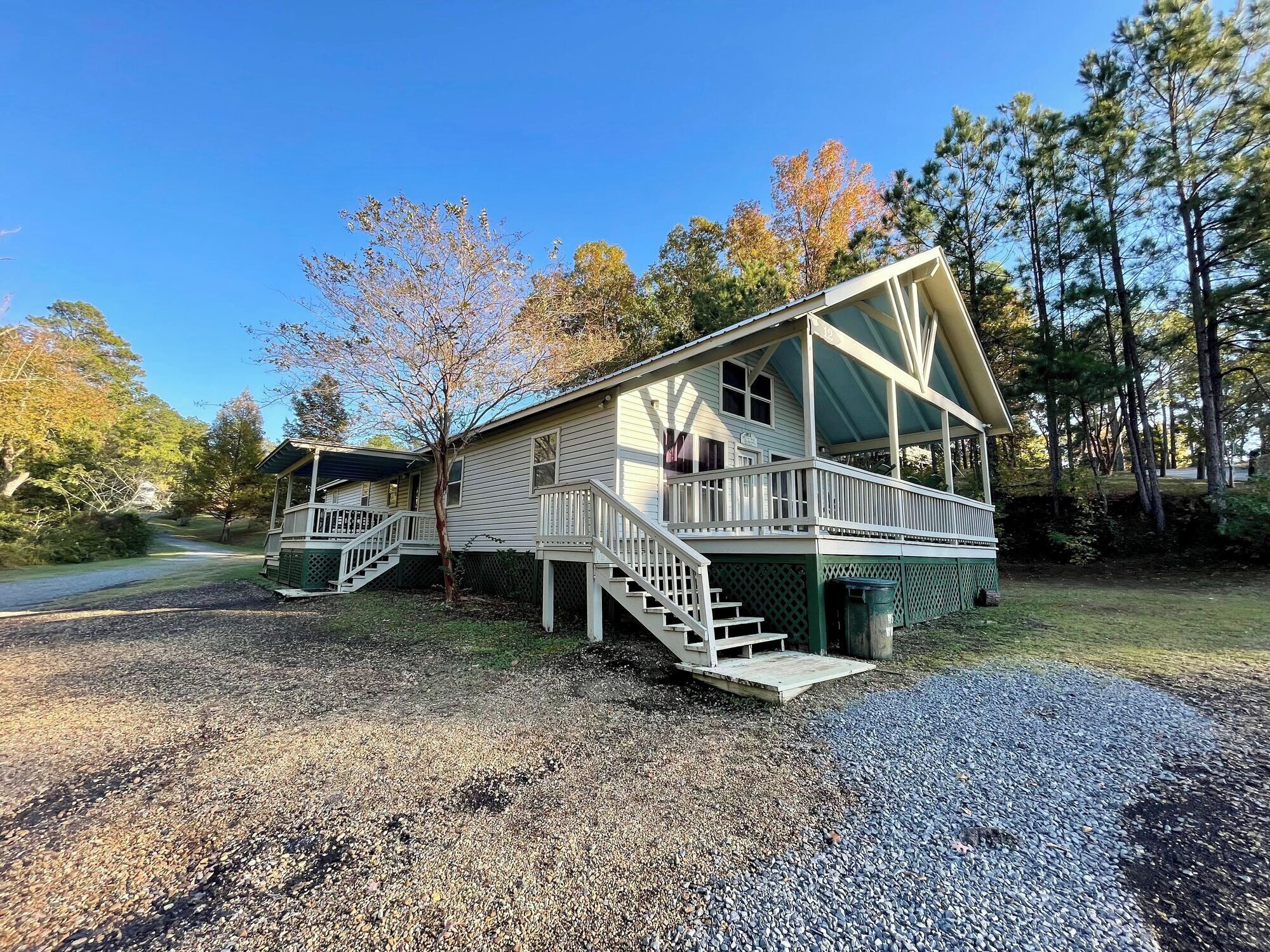 A two-story cabin with a porch and stairs, surrounded by trees and gravel.