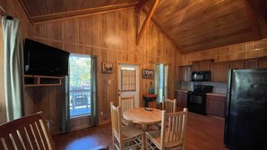 A wooden interior with a dining table, chairs, a TV, and a kitchen area including a microwave and refrigerator. - Peaceful Cabin with Full Kitchen, Vaulted Ceilings & Pond View Porch (Zwolle)