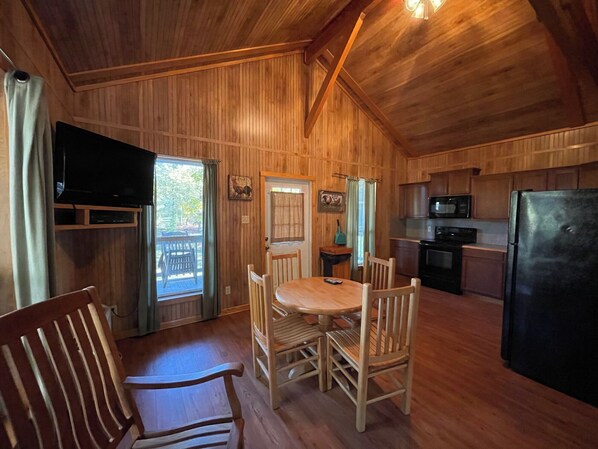 A wooden interior with a dining table, chairs, a TV, and a kitchen area including a microwave and refrigerator. - Peaceful Cabin with Full Kitchen, Vaulted Ceilings & Pond View Porch (Zwolle)
