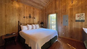 A wooden room with a bed, a nightstand, a wall-mounted light fixture, a window with curtains, and a sign on the wall. - Peaceful Cabin with Full Kitchen, Vaulted Ceilings & Pond View Porch (Zwolle)