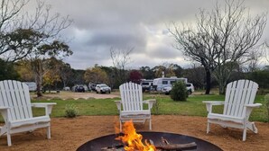 A campfire with two white Adirondack chairs facing it, set on a gravel area with trees and RVs in the background. - Country Style Holiday Park (Glen Aplin)