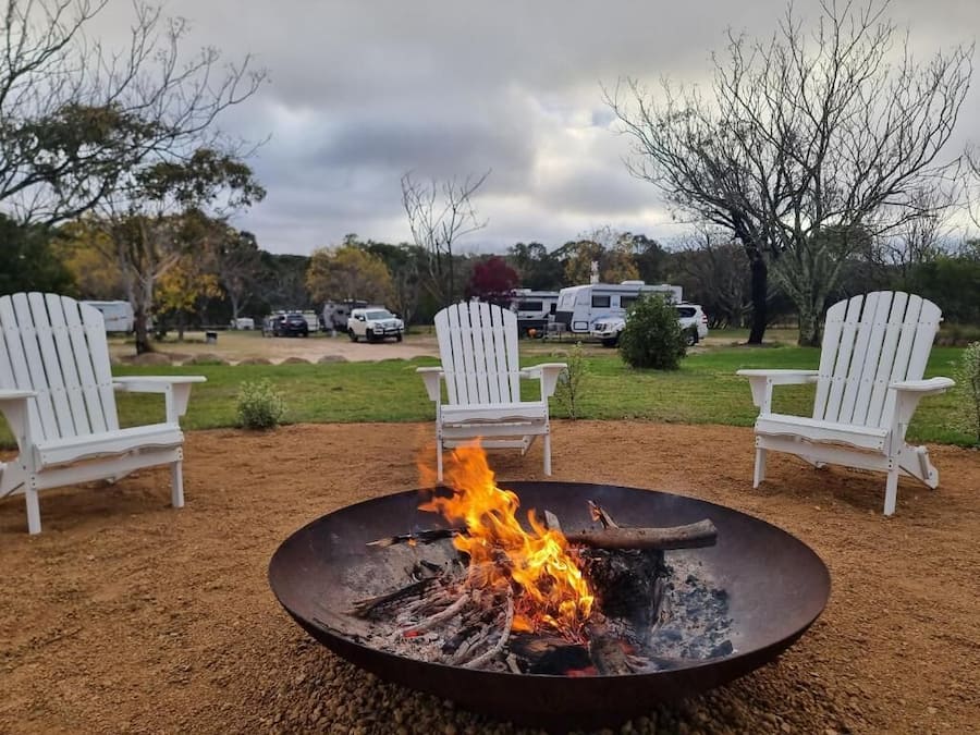 A campfire with two white Adirondack chairs facing it, set on a gravel area with trees and RVs in the background.