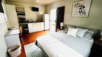 A compact hotel room with a bed, bedside table, lamp, and a kitchenette area.