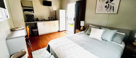 A compact hotel room with a bed, bedside table, lamp, and a kitchenette area.