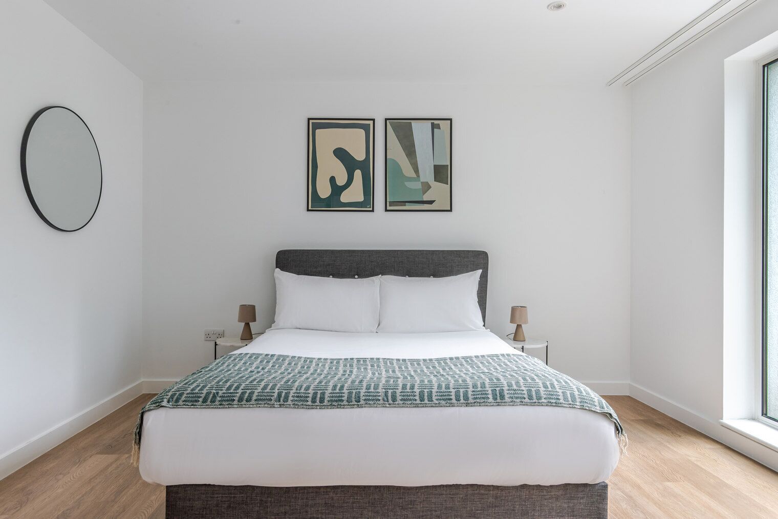A modern bedroom with a large bed, two bedside tables, a round mirror, and abstract artwork on the wall.