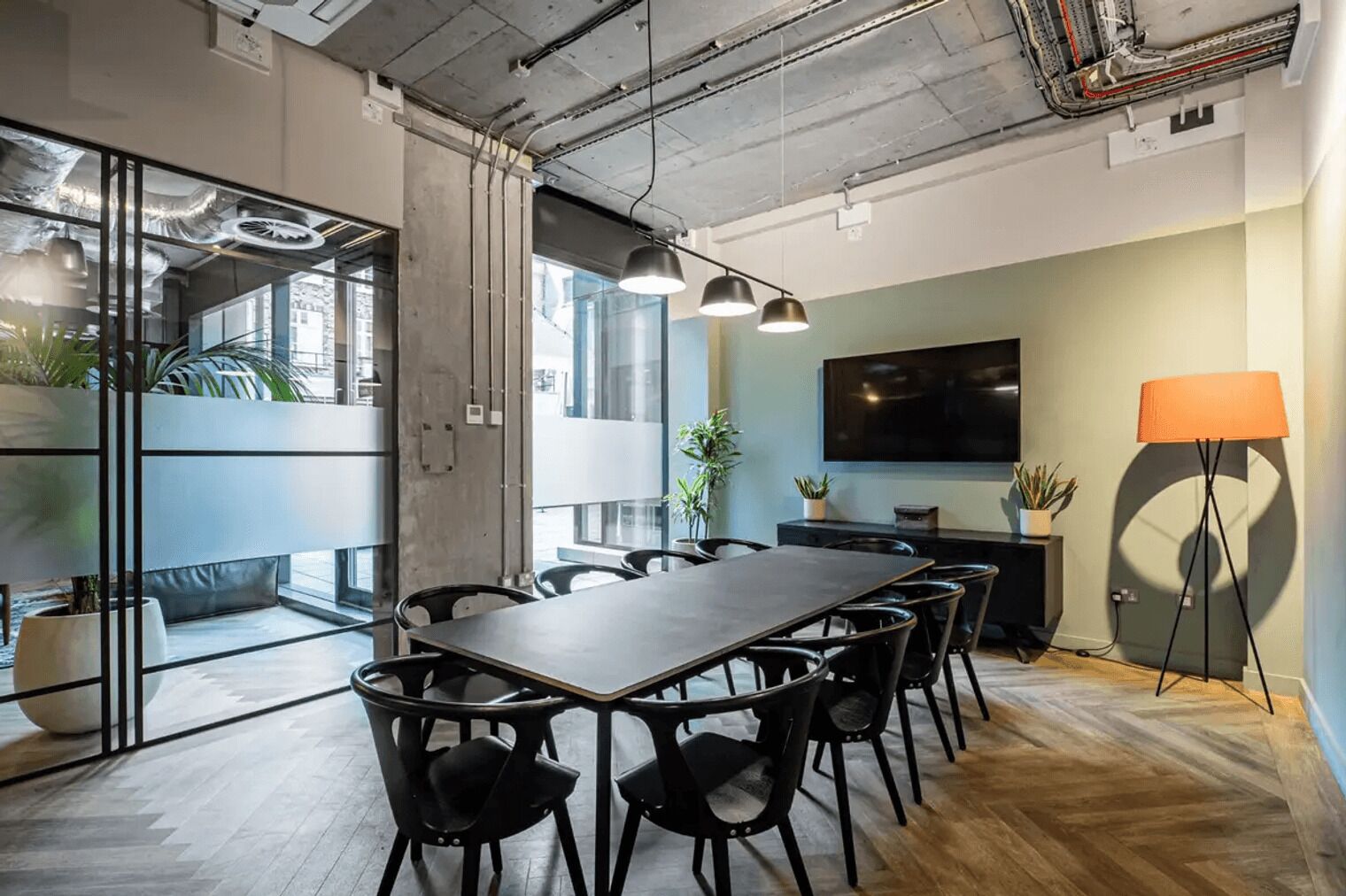 A modern conference room with a long table, black chairs, a large TV, and a floor lamp.