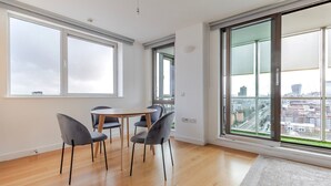 Een moderne woonkamer met een houten vloer, een ronde tafel en vier grijze stoelen. Grote ramen bieden uitzicht op een stadsgezicht.