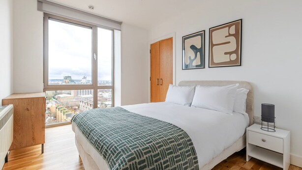 Een moderne slaapkamer met een groot raam, een bed met een geborduurd sprei, een nachtkastje en twee abstracte kunstwerken aan de muur.