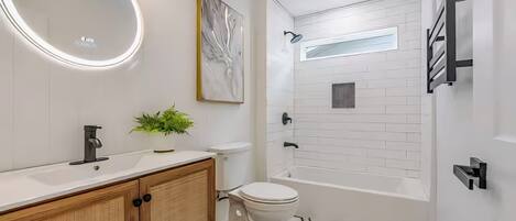 Une salle de bain moderne avec un lavabo blanc, un meuble-lavabo en bois, un miroir rond, une plante en pot, des toilettes et une baignoire dont le sol est recouvert de carreaux aux motifs géométriques.