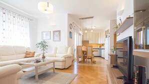 A modern living room with a sofa, coffee table, and TV. There is a kitchen in the background with cabinets and a dining area. - Holiday apartment with balcony (Biograd na Moru)