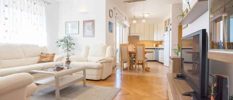 Un salon moderne avec un canapé, une table basse et un téléviseur. On aperçoit au fond une cuisine équipée de placards et un coin repas.