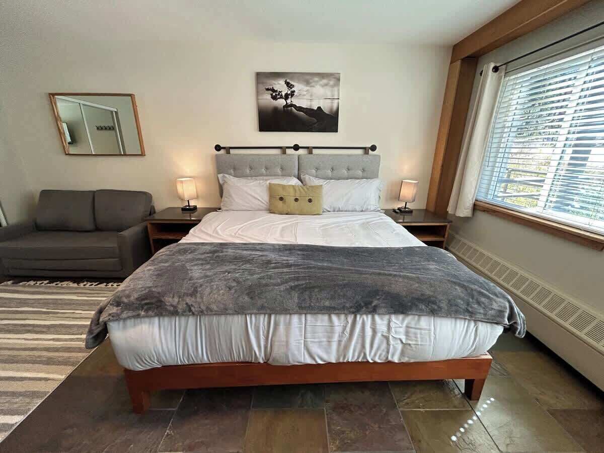A bedroom with a large bed, a nightstand, a bedside table, a sofa, a painting on the wall, and a window with blinds.