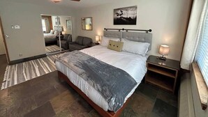 Una habitación de hotel moderna con una cama grande, un ventilador de techo, una mesita de noche con lámpara, un espejo y un cuadro en la pared.