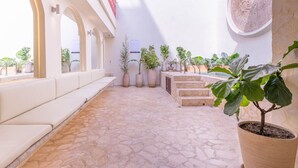 Un patio luminoso con suelo de piedra, asientos blancos y plantas en macetas.
