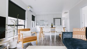Un salon moderne avec un canapé bleu foncé, une chaise en bois et un fauteuil blanc. On y trouve également un coin repas avec des chaises blanches et une table. La cuisine est équipée de meubles blancs et d’un réfrigérateur.
