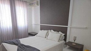 Uma cama bem arrumada com um cobertor cinzento, uma mesinha de cabeceira com um objeto decorativo e uma cadeira.
