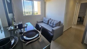 Uma sala de estar moderna com um sofá cinzento, uma mesa de jantar com cadeiras e uma janela grande que oferece vista para o exterior.