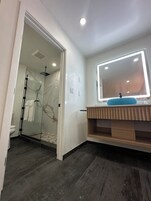 Un baño moderno con ducha envuelta en vidrio, tocador con lavabo azul y un espejo grande con iluminación LED.