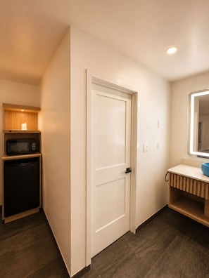 A compact room with a white door, a small kitchenette with a microwave and a refrigerator, and a mirror above a sink. - Nubell Motel (Bell Gardens)