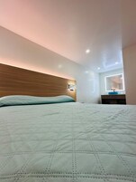 Una cama bien hecha con cubrecama acolchado, cabecera de madera, mesita de noche con lámpara y espejo con luces.