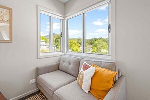 Ein Eckzimmer mit grauem Sofa, zwei Fenstern und Blick auf Grünflächen und Gebäude.