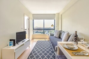 Ein modernes Wohnzimmer mit großem Fenster, einem Flachbildfernseher, einer Couch und einem Essbereich mit Tisch für zwei Personen.