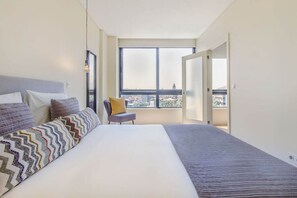 Ein Schlafzimmer mit einem großen Bett, einem Sessel und Blick auf die Stadt durch ein großes Fenster.