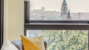Une chambre avec un lit, une fenêtre donnant sur un paysage urbain et un beffroi, ainsi qu’un luminaire suspendu.