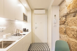 Eine schmale Küche mit integriertem Mikrowellenherd, Wasserkocher und Kaffeemaschine. Ein Spülbecken mit modernem Wasserhahn, darüber ein kleines Fenster. Der Boden ist mit einem geometrischen Fliesenmuster versehen.