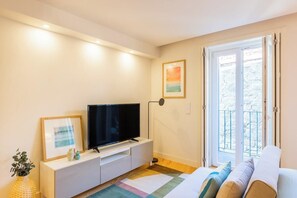 Een moderne woonkamer met een flatscreen televisie op een witte console, een ingelijst beeld, een vloerlamp, een kleurrijk vloerkleed en een balkon met uitzicht.