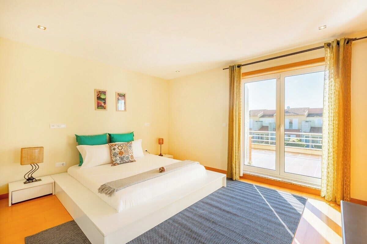 Une chambre à coucher avec un grand lit, une table de chevet, une lampe et une vue sur les immeubles par la fenêtre.