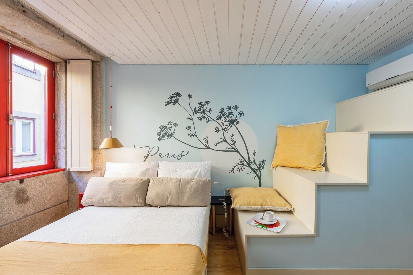A bedroom with a bed, pillows, a wall-mounted shelf with a cup and saucer, a window with a red frame, and a wall decal with the word "Paris".