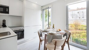 Eine moderne Küche mit weißen Schränken, einem eingebauten Backofen und einer Mikrowelle. Es gibt eine Essecke mit einem Tisch für zwei Personen und ein großes Fenster mit Blick auf einen Garten und Gebäude.