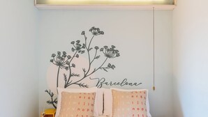 Ein Schlafzimmer mit einem Bett, zwei Kissen mit alphabetischen Mustern, einer Wanddekoration mit Blumen und dem Schriftzug 'Barcelona' sowie einem roten Hocker.