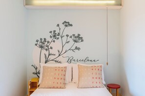 Ein Schlafzimmer mit einem Bett, zwei Kissen mit alphabetischen Mustern, einer Wanddekoration mit Blumen und dem Schriftzug 'Barcelona' sowie einem roten Hocker.