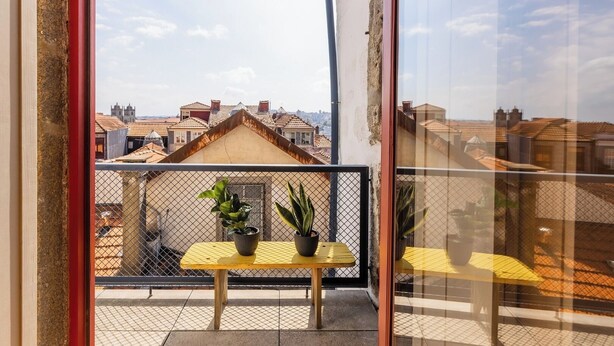 Ein Balkon mit einem gelben Tisch und zwei Blumentöpfen, mit Blick auf eine Stadtlandschaft mit traditionellen Dächern.